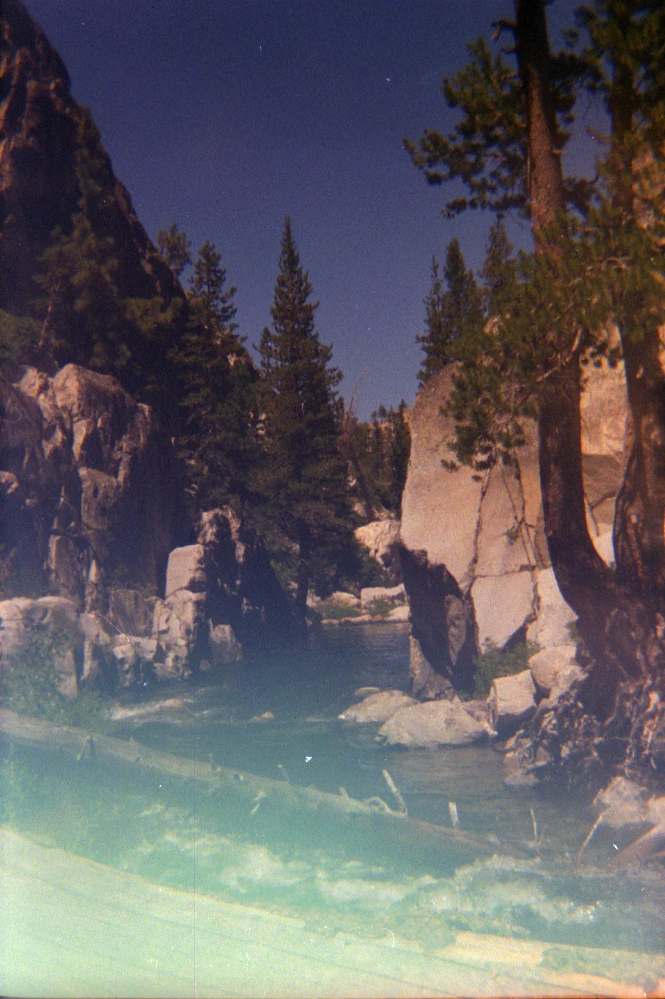 Rocky river gorge with boulders, pine trees, and rushing water under a blue sky, slightly faded vintage tones