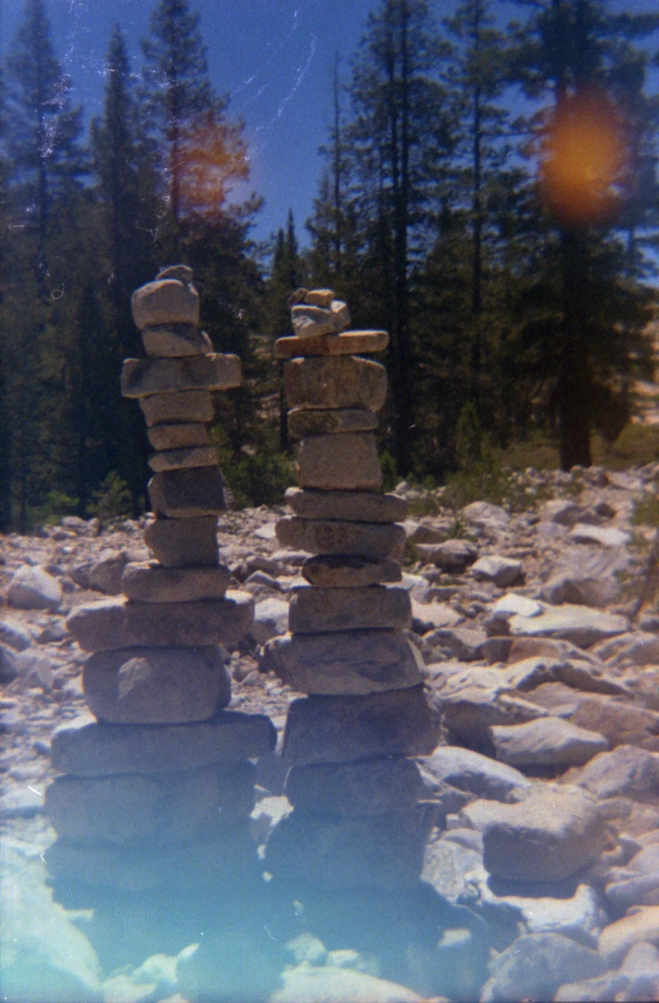 Two stacked stone cairns on a rocky trail surrounded by pine trees under a blue sky, with sun flare, natural serene mood